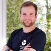 Physiotherapeut Niklas Witt trägt ein schwarzes T-Shirt und lächelt in die Kamera. Im Hintergrund grüne Bäume.